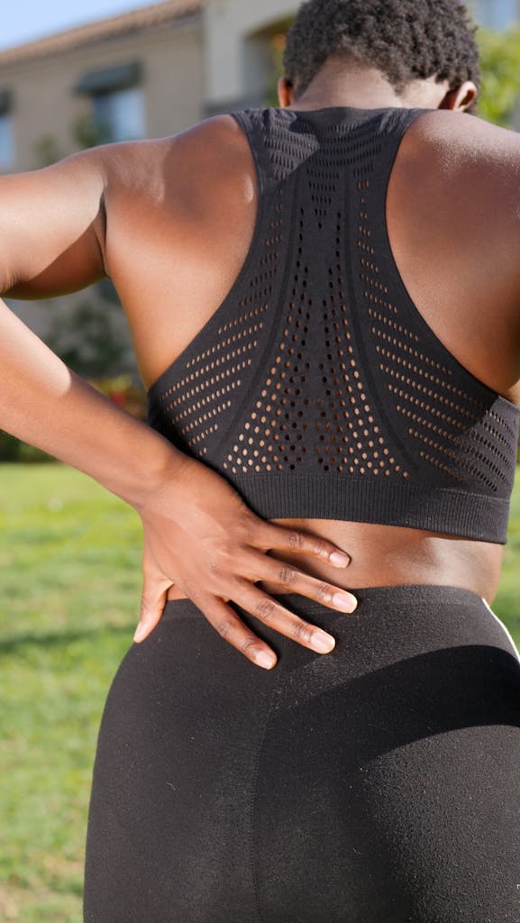 A woman in sportswear gripping her lower back, potentially indicating discomfort, set in an outdoor environment.